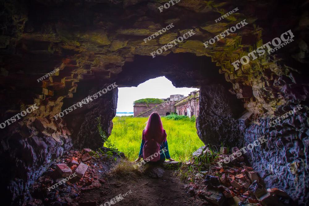 На фотографии изображена девушка, сидящая под аркой в старинной крепости. За аркой открывается вид на зелёные луга и стены разрушенной крепости. Этот снимок передаёт ощущение покоя и задумчивости на фоне исторической архитектуры.